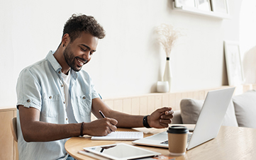 Young male sitting and smiling at his desk while writing notes on paper